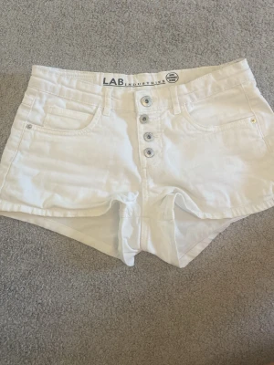 Vita low waist shorts med knappar - Snygga vita shorts med låg midja och fyra synliga knappar framtill. Klassisk femficksmodell i bomullsmix, perfekta för varma dagar. Passar till allt från crop tops till hoodies och ger en fräsch look. Säljer eftersom den inte kommer till användning