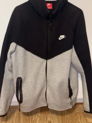 Grå och svart Nike tech fleece - Nike hoodie i svart och grått med dragkedja framtill och två fickor med dragkedja. Klassisk Nike-logga på bröstet och huva med snörning. Tillverkad i mjukt material som passar perfekt för en avslappnad streetwear-look.