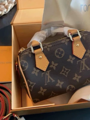 Monogram handväska i brunt skinn - Snygg handväska i brunt skinn med klassiskt monogrammönster och detaljer i beige läder. Väskan har guldfärgade metalldetaljer och dragkedja upptill. Perfekt storlek för det viktigaste och en ikonisk accessoar till din outfit.