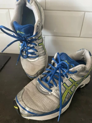 Grå-blå sneakers med gula detaljer - Snygga sneakers i grått med blå och gula detaljer samt blå snörning. Ovandelen är i mesh och syntetmaterial för bra andningsförmåga. Sulan har svarta och gula inslag och skon har en sportig vibe. Perfekta för dig som gillar en aktiv stil. Nytvättade 