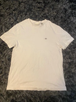 Vit basic t-shirt med liten logga - En enkel vit t-shirt i bomull med rund hals och korta ärmar. På bröstet finns en liten diskret logga som ger en clean look. Perfekt till jeans eller shorts för en avslappnad stil.Använd fåtal gånger gant tshirt (äkta) den har en jätteliten fläck baksidan av tshirten som ni ser i sista bilden jag tror ochså den går bort inget så farligt annars inga skador eller nått den är i size S och är i mycket bra skick(passar även M)