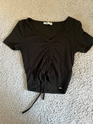 Svart ribbad topp med snörning - Svart ribbad topp från FB Sister med v-ringning och kort ärm. Toppen har snörning framtill som ger en snygg rynkad effekt och croppad passform. Perfekt att styla med jeans eller kjol för en trendig look.
