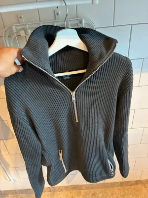 Filippa K - Quarterzip - L - Mörkgrå ribbstickad tröja med hög krage och silverfärgade dragkedjor framtill samt vid fickorna. Tröjan har lång ärm och en modern, avslappnad passform. Perfekt för kyliga dagar och enkel att matcha med olika outfits.