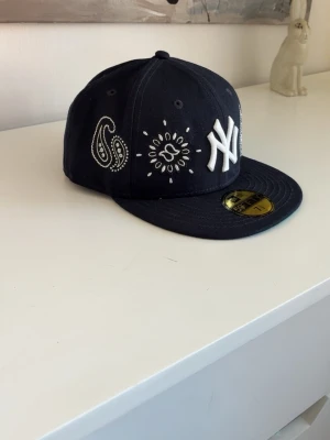 Blå  New Era NY keps med brodyr - Snygg blå  keps från New Era med New York Yankees-logga framtill. Kepsen har vit brodyr i paisleymönster och andra detaljer runt loggan. Flat skärm och klassisk 59FIFTY-modell. Perfekt för dig som gillar streetwear och vill sticka ut.