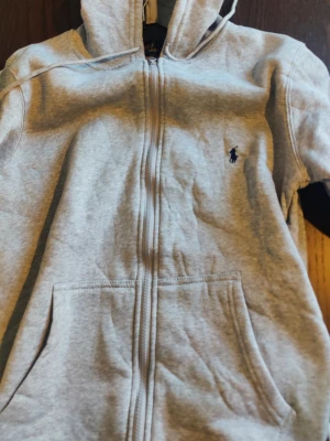 Grå zip hoodie från Ralph Lauren - Snygg grå hoodie med dragkedja från Polo Ralph Lauren. Klassisk design med huva, kängurufickor och den ikoniska mörkblå loggan broderad på bröstet. Tillverkad i mjukt bomullsmaterial som är perfekt för chill dagar.