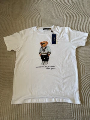 Vit Polo Bear t-shirt från Ralph Lauren - Vit t-shirt från Ralph Lauren med ikoniskt Polo Bear-tryck framtill. Klassisk passform med rund hals och korta ärmar. Tillverkad i mjuk bomull som känns skön mot huden. Perfekt för en avslappnad och trendig look.