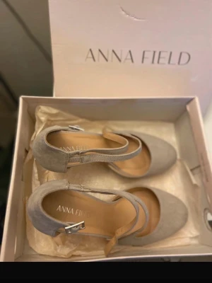 Beige klackar med rem från Anna Field - Snygga beige pumps från Anna Field i mockaimitation med rund tå och justerbar rem med silverfärgat spänne. Klacken är medelhög och skon har en klassisk, stilren design som passar till många olika outfits.