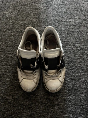 Valentino opens - Säljer ett par vita sneakers med bred svart rem över vristen och klassisk snörning. Skorna har en låg silhuett och är tillverkade i läder eller konstläder. Perfekta för en avslappnad streetwear-look.