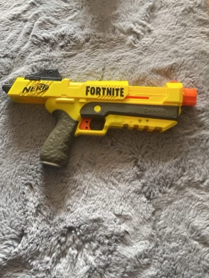 Nerf Fortnite gul leksakspistol - Cool gul Nerf Fortnite-leksakspistol i plast med detaljer i svart, orange och grått. Pistolen har Fortnite-logga på sidan och ett greppvänligt handtag. Perfekt för cosplay eller maskerad som Fortnite-karaktär.