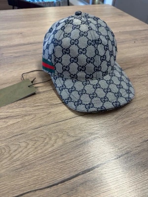 Gucci keps med GG-mönster - Snygg keps från Gucci i grått tyg med klassiskt GG-mönster i mörkblått. Kepsen har böjd skärm och en grön-röd rand på sidan som ger en ikonisk touch. Perfekt accessoar för att lyfta din streetwear-look.