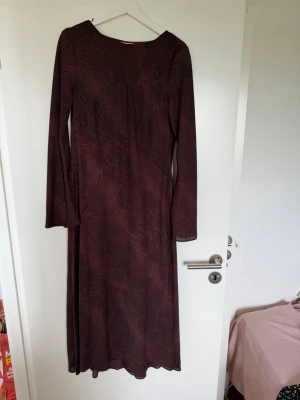 Mörkröd långklänning med mönster - Långklänning i mörkröd ton med diskret mönster. Klänningen har lång ärm och rak passform, perfekt för dig som gillar en stilren look. 