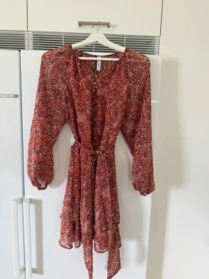 Blommig kortklänning från MNG - Supersöt kortklänning från MNG med blommigt mönster i rött, orange och vitt. Klänningen har långa ärmar, knappar framtill och ett matchande bälte i midjan. Perfekt för vår och sommar med sitt lätta, luftiga material.