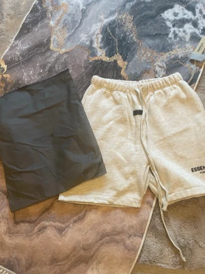 Beige Essentials shorts med snörning - Säljer ett par ljusbeige shorts från Essentials Fear of God. De har elastisk midja med långa snören framtill och logga tryckt på vänster ben. Perfekta för chill dagar och har en relaxed fit.