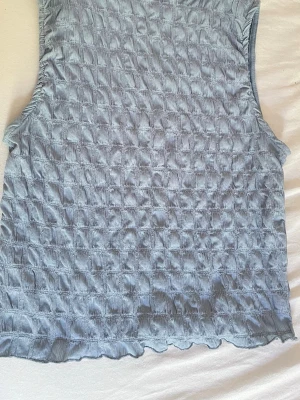 Ljusblå ribbad ärmlös linnetopp - Säljer en ljusblå ärmlös linnetopp med ribbad struktur och vågig kant nertill. Toppen har en stretchig och texturerad yta som ger en cool och trendig look. Perfekt att styla med jeans eller kjol för en avslappnad vibe.