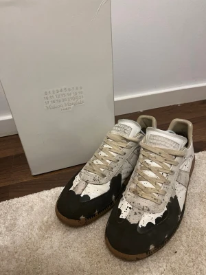 Maison Margiela paintsplater  - Unika sneakers från Maison Margiela i vitt och beige med svart färgstänksdesign och gummisula. Lågt skaft och klassisk siluett med snörning. Tillverkade i läder och textil för en cool och trendig look.