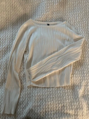 Vit ribbad långärmad topp - Säljer en vit långärmad topp med ribbade vågiga detaljer. Toppen har en något croppad passform och är tillverkad i ett mjukt, stretchigt material som känns skönt mot huden. Perfekt att styla med jeans eller kjol för en clean look.