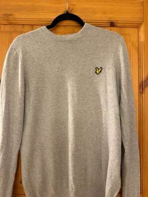 Ljusgrå stickad tröja med logga - Ljusgrå stickad tröja med rund hals och lång ärm. På bröstet finns en broderad gul och svart logga i form av en fågel. Tröjan har ribbade muddar vid ärmslut och nederkant. Perfekt för lager-på-lager och enkel att matcha.