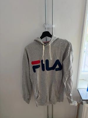 Grå hoodie från FILA med logga - Snygg grå hoodie från FILA med stor logga i rött och blått framtill. Hoodien har huva med vita dragsnören och en klassisk känguruficka. Perfekt för en avslappnad och sportig stil.