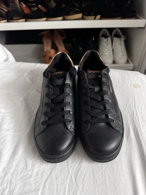 Svarta sneakers från Björn Borg - Stilrena svarta sneakers från Björn Borg i läderimitation med svarta skosnören och en snygg detalj i metallic guld på hälen. Klassisk låg modell med diskret logga på sidan och plös. Perfekta för en clean och modern look.