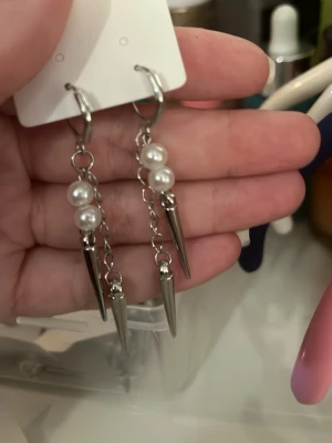 Silvriga örhängen med pärlor och spikes - Säljer ett par statement örhängen i silverfärg med kedjor, vita pärlor och coola metallspikar. Örhängena har en edgy vibe och passar perfekt för dig som vill sticka ut med din stil. Säljs som par.