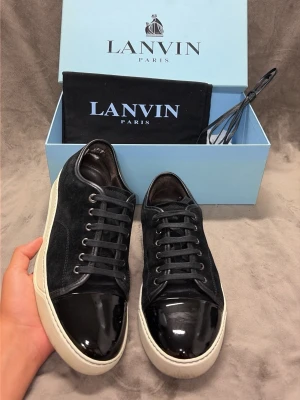 Svarta Lanvin sneakers med lackad tå - Snygga svarta sneakers från Lanvin med ovandel i mocka och en glansig, lackad tå. Skorna har svarta skosnören och vit sula som ger en clean kontrast. Perfekta för dig som vill ha en stilren och lyxig look.