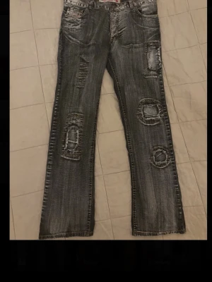 Grå slitna jeans med patchwork - Säljer ett par grå jeans med coola slitningar och patchwork-detaljer på benen. Jeansen har raka ben och snygga bakfickor med mönster. Perfekta för dig som gillar en edgy och unik stil. Materialet är klassisk denim.