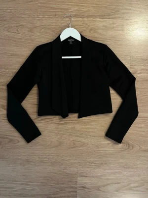 Svart kort kavaj från Boohoo - Stilren svart kavaj från Boohoo med öppen front och croppad passform. Långa ärmar och mjukt, stretchigt material som ger en bekväm känsla. Perfekt för att lyfta vilken outfit som helst med en modern touch.