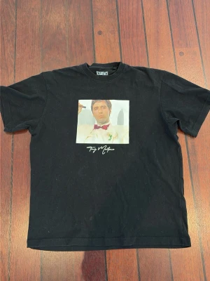 Svart Scarface t-shirt med tryck - Cool svart t-shirt med stort Scarface-tryck på ryggen och motiv av Tony Montana på framsidan. Klassisk passform och rund hals. Perfekt för dig som gillar film och streetwear.