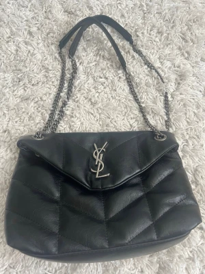 Svart handväska från Saint Laurent - Snygg svart quiltad handväska från Saint Laurent med silverfärgad YSL-logga framtill. Väskan har kedjeremmar med läderdetaljer och ett rymligt innerfack med dragkedja. Tillverkad i mjukt läder och har en klassisk, tidlös design.