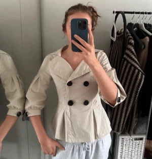 Cremefärgad trenchcoat med stora knappar - Stilren beige trenchcoat med dubbelknäppning och stora svarta knappar. Storlek L men mer som en S/M