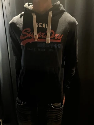  Superdry hoodie - Svart hoodie från Superdry med stor logga i rött, vitt och blått framtill. Tröjan har huva med vita dragsnören och en känguruficka. Perfekt för dig som gillar vintagestilen och vill ha en skön och snygg hoodie 👊