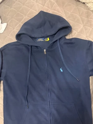 Marinblå hoodie från Polo Ralph Lauren - Säljer en marinblå hoodie från Polo Ralph Lauren med dragkedja framtill och klassisk huva med dragsko. Ikoniska blå loggan broderad på bröstet. Tillverkad i mjukt bomullsmaterial som är skönt att bära. Perfekt för en avslappnad och stilren look. SIZE M PASSAR SOM S