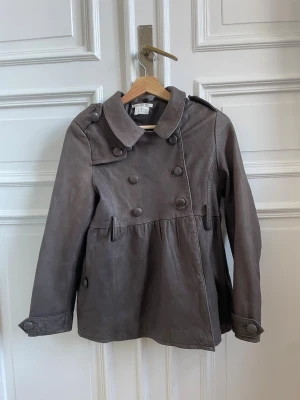 Brun trenchcoat med dubbelknäppning - Snygg brun trenchcoat med dubbelknäppning och stora dekorativa knappar. Jättefin och bra material! 