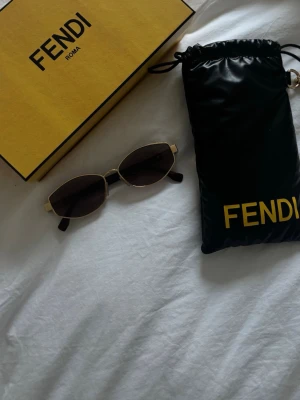 Fendi svarta solglasögon med gulddetaljer - Snygga solglasögon från Fendi med svarta glas och tunna guldfärgade bågar. Ovala linser och stilren design som ger en lyxig känsla. Kommer med originalfodral och ask. Perfekt accessoar för att lyfta din look. Köpta för 4300kr, 2026 model