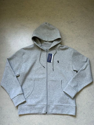 Grå hoodie från Polo Ralph Lauren - Ralph Lauren zip-tröja – Grå – Storlek M  Helt ny och oanvänd Ralph Lauren zip-tröja i grått. Bekväm passform och hög kvalitet. Perfekt till både vardag och en stilren look.  • Storlek M • Helt ny och oanvänd • Dragkedja (zip) • Grå färg • Bekväm och skön
