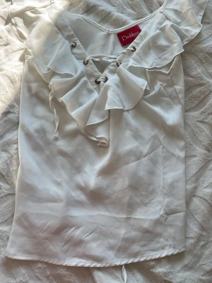 Vit blus med volang och snörning - Supersöt vit blus från Diabless med volangdetaljer längs ringningen och snörning framtill. Blusen är ärmlös och har en lätt, flowy passform som känns luftig och somrig. Perfekt att styla med jeans eller kjol för en fräsch look.