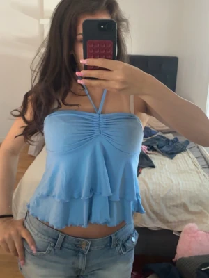 Ljusblå volang topp med halterneck -  ljusblå topp med halterneck och dubbla volanger. Toppen är ärmlös och har en rynkad detalj framtill som ger en snygg form. Perfekt till jeans eller kjol 