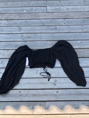Svart lacing smock blus från Nelly - Trendig svart blus från Nelly med puffiga långärmar och snörning framtill. Modellen är croppad och har en smockad design som ger en snygg siluett. Perfekt för dig som vill ha en statement-blus med coola detaljer.