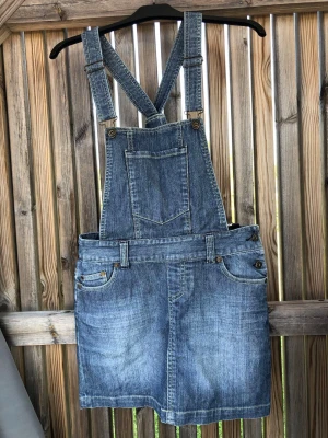 Jeansskort med hängslen i blå denim - Snygg jeansskort i klassisk blå denim med justerbara hängslen och bröstficka framtill. Skorten har fickor både fram och bak samt bälteshällor. Perfekt för en avslappnad och trendig look.