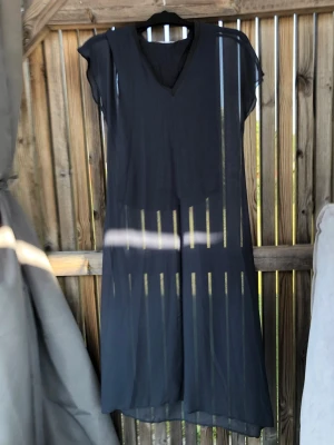Svart transparent strandklänning - Snygg svart strandklänning i transparent, lätt material. Klänningen har korta ärmar, v-ringning fram och rak passform. Perfekt att dra över bikinin på stranden för en avslappnad och trendig look.