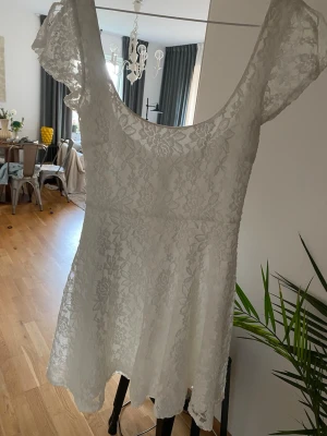 Vit spetsklänning med kort ärm - Söt vit kortklänning i spets med blommigt mönster och rundad urringning både fram och bak. Klänningen har korta ärmar och en lätt utställd kjol som ger en romantisk känsla. Perfekt för sommarens alla tillfällen.