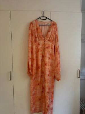 Adoore klänning - Säljer en lång klänning i ljusa rosa och orange toner med blommigt mönster. Klänningen har v-ringning, långa puffärmar och knappar framtill. Perfekt för sommarens alla tillfällen och ger en romantisk vibe.
