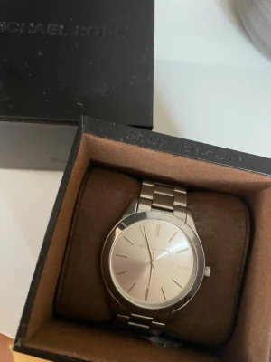 Silverfärgad klocka från Michael Kors - Stilren klocka från Michael Kors i silverfärg med metallarmband och minimalistisk urtavla. Klockan har en rund form och klassisk design utan siffror, vilket ger en clean och modern look. Perfekt accessoar för dig som gillar enkelhet och elegans.