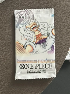 OP-05 One Piece kortspel boosterpack  - Säljer ett boosterpack till One Piece Card Game med temat Awakening of the New Era. Förpackningen är vit med färgglad animeillustration och innehåller 10 extra spelkort. Perfekt för samlare eller fans av One Piece.