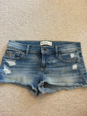 Blå jeansshorts från Abercrombie - Säljer ett par blå jeansshorts från Abercrombie med slitna detaljer och fransig kant. Klassisk femficksmodell med låg midja och snygga slitningar både fram och bak. Perfekta för varma dagar och en avslappnad stil.💓