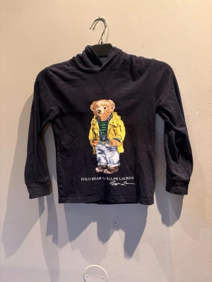 Svart hoodie med Polo Bear-tryck - Svart hoodie med lång ärm och huva från Ralph Lauren. På bröstet finns ett stort färgglatt tryck av den ikoniska Polo Bear i gul jacka, randig tröja och jeans. Under motivet står texten 'POLO BEAR by RALPH LAUREN'. Perfekt för en avslappnad och cool stil.