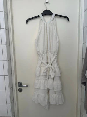 Vit ärmlös klänning med volanger - Supersöt vit klänning med halterneck, knytband i midjan och volangdetaljer på benen. Tunt, luftigt tyg med diskret strukturmönster. Perfekt för sommarens alla äventyr och ger en riktigt chill vibe.