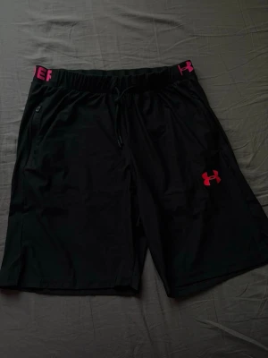 Svarta träningsshorts Under Armour - Säljer ett par svarta träningsshorts från Under Armour med rosa logga och resår i midjan. Shortsen har dragsko och elastisk midja med texten 'Under Armour' i rosa. Perfekta för gym eller löpning.