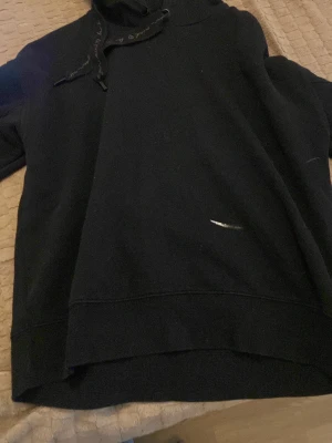 Svart hoodie med huva och snören - En enkel svart hoodie med huva och matchande svarta snören. Hoodien har en rak passform och ribbade muddar vid ärmslut och nederkant. Perfekt för en avslappnad stil och funkar året runt. Materialet ser ut att vara mjukt och bekvämt, troligen bomull eller bomullsblandning.