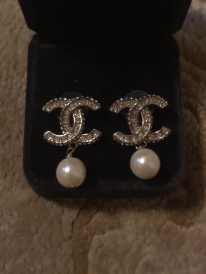 Chnel örhängen med pärldetalj - Säljer ett par ikoniska Chnel örhängen i silverfärg med gnistrande stenar i det klassiska CC-mönstret. Under hänger en elegant vit pärla som ger en lyxig touch. Perfekta för dig som vill ha något extra till din stil.